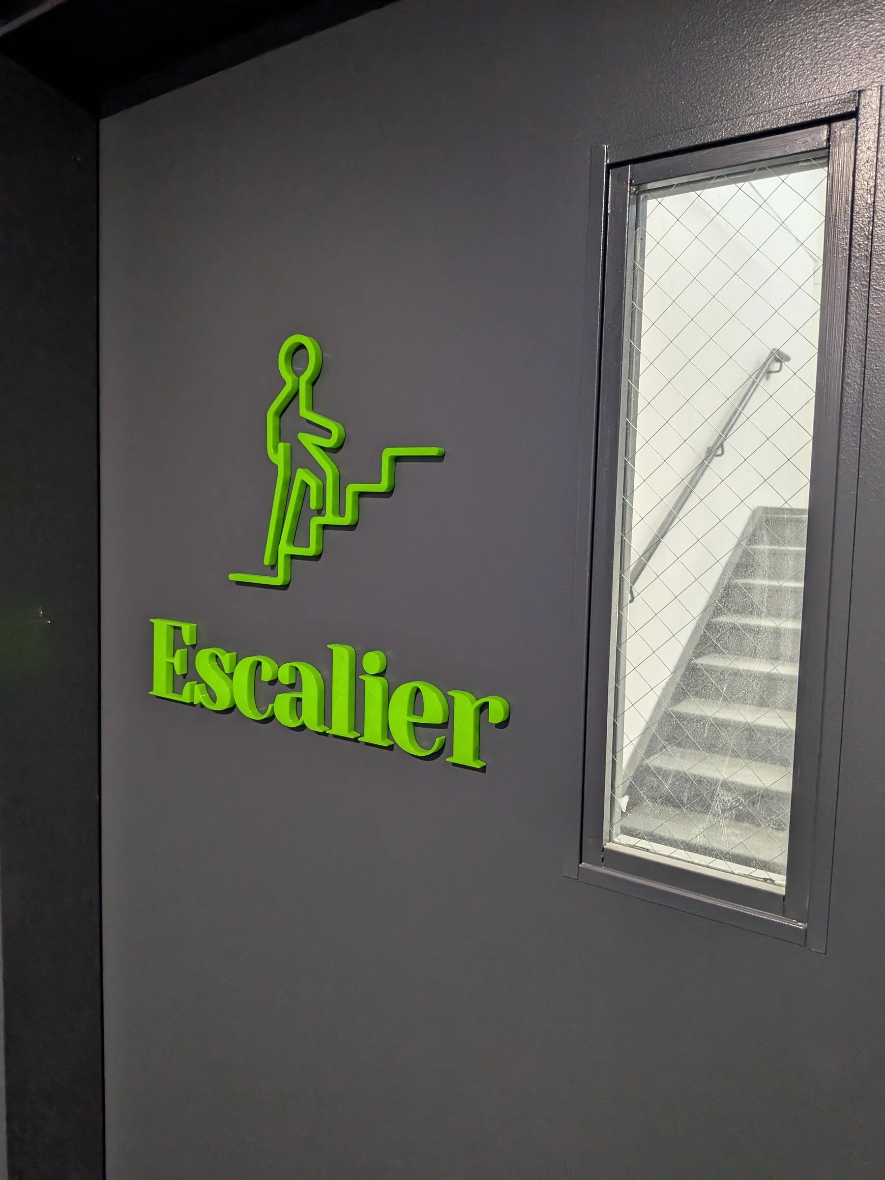 Pictogramme 3D « Escalier » vert et noir posé sur un mur gris, Cinq23