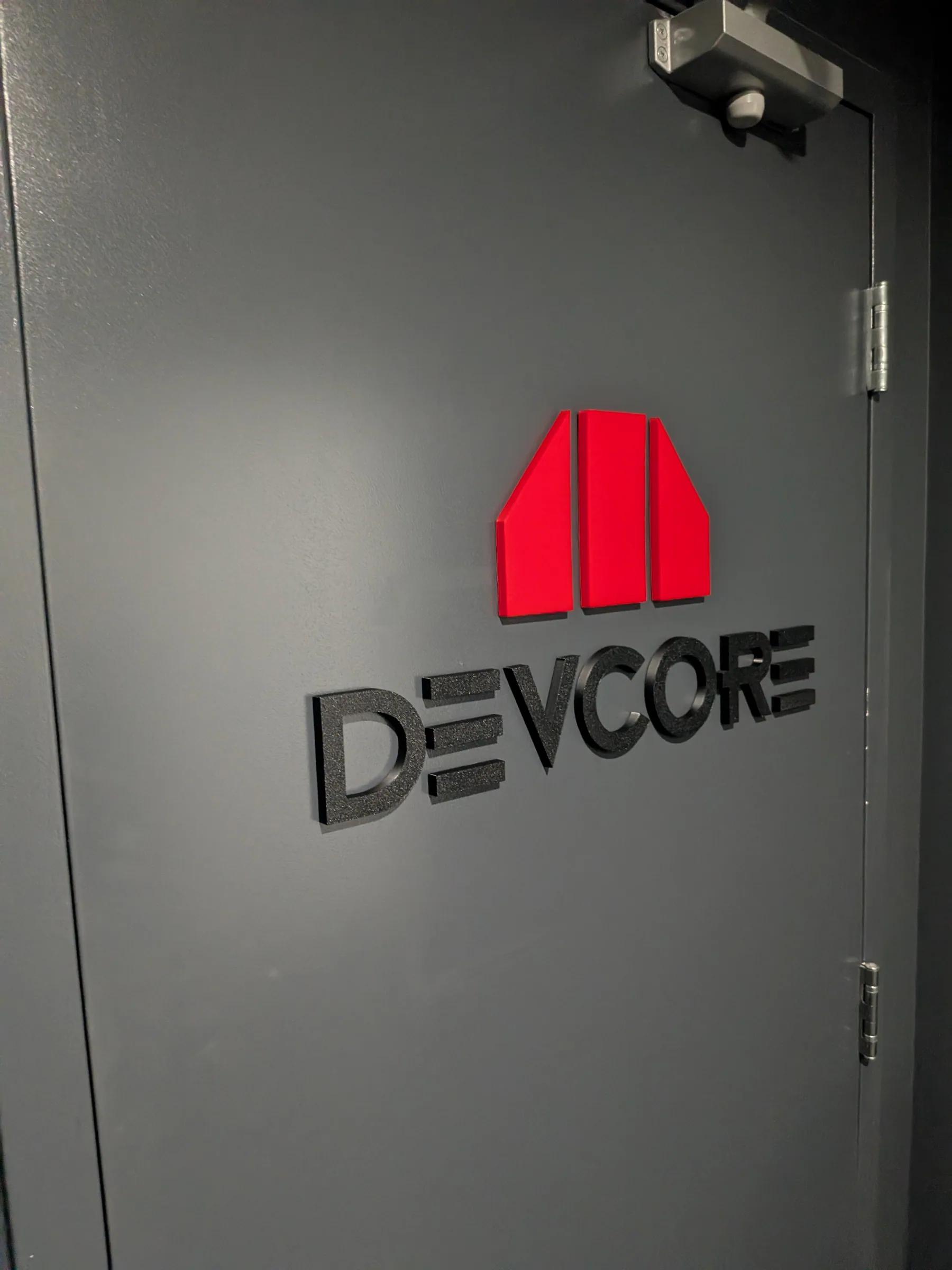 Enseigne Devcore en lettres 3D noires et logo rouge sur un mur gris