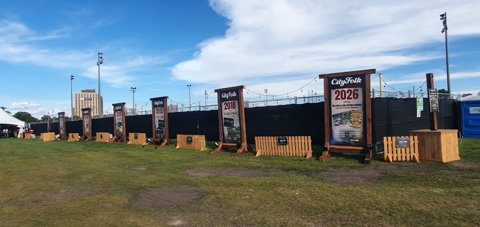 Panneaux CityFolk 2011, 2015, 2018 et 2026 sur structures de bois, barrières de bois au pied, pelouse du festival
