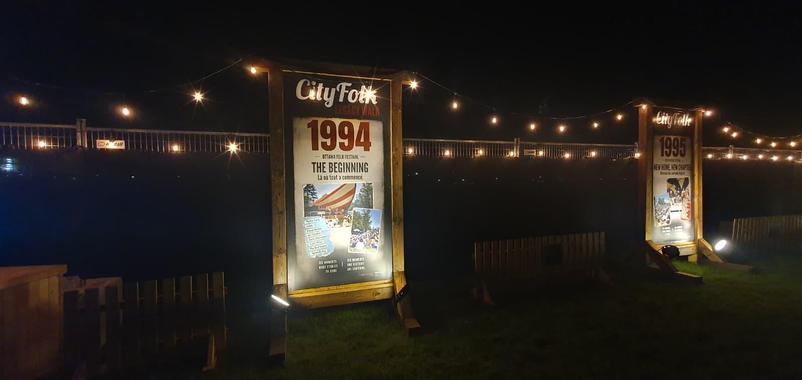 Panneau CityFolk 1994 « The Beginning » éclairé de nuit, guirlandes lumineuses tendues derrière