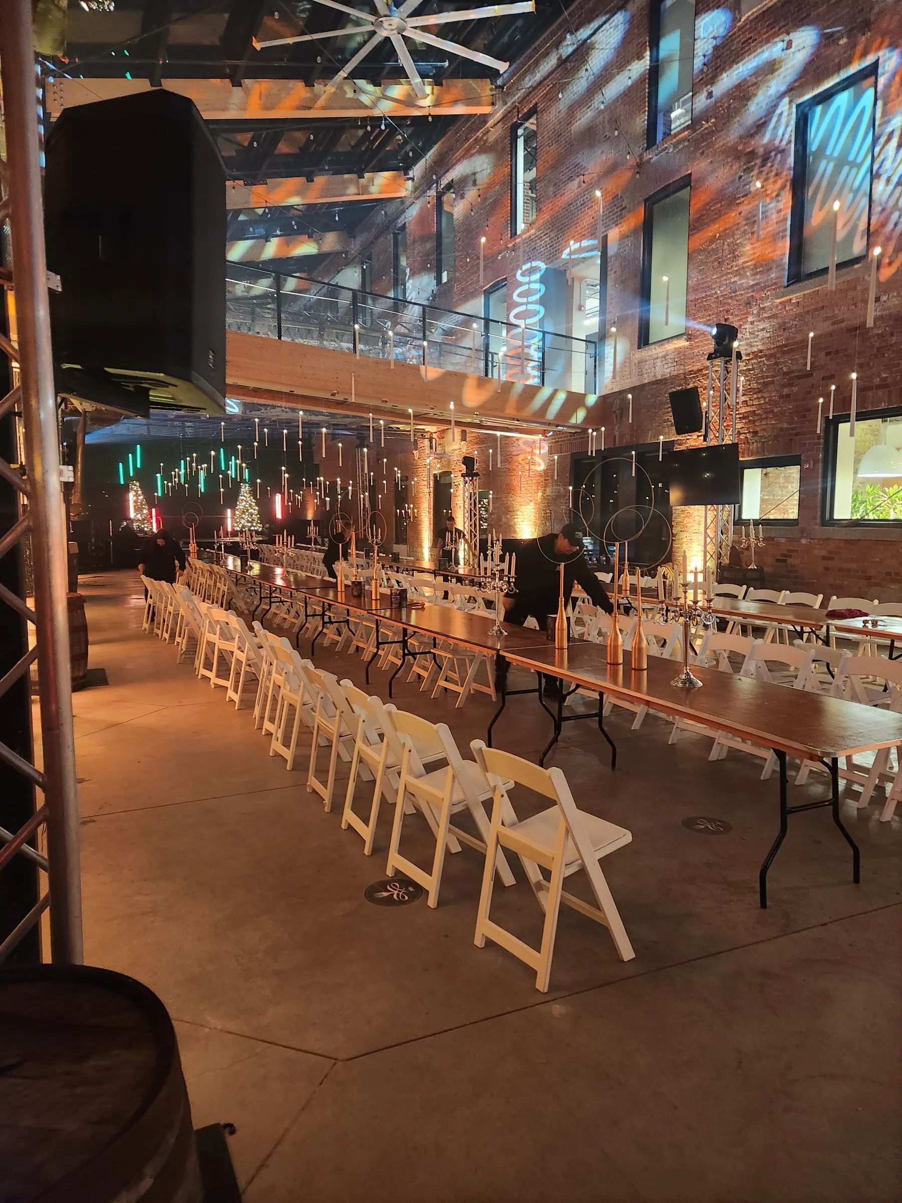Longues tables dressées, chaises blanches et chandeliers dans une salle en brique